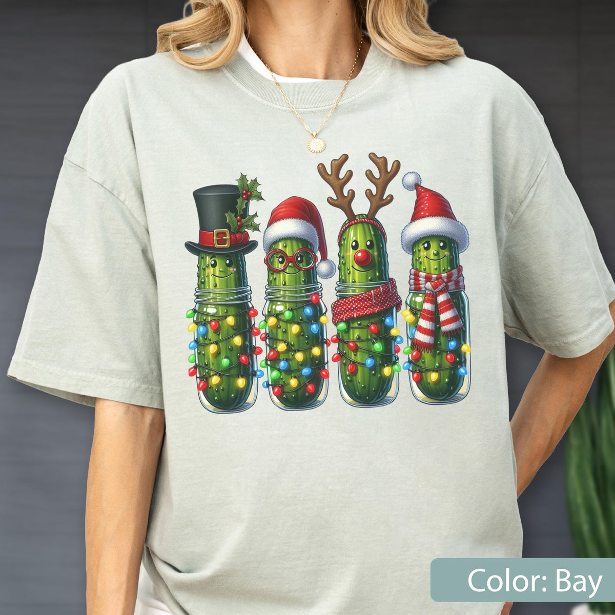 May include: A light gray t-shirt featuring a graphic of four green pickles in glass jars. Each pickle is decorated for the holidays with items including a top hat, Santa hat, reindeer antlers, a red nose, scarves, and colorful string lights.
