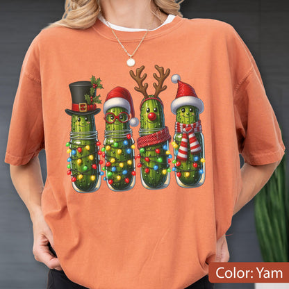 May include: A burnt orange short-sleeve t-shirt featuring a graphic of four green pickles decorated for the holidays. The pickles wear festive accessories including a top hat, Santa hats, reindeer antlers, a red nose, and colorful string lights.