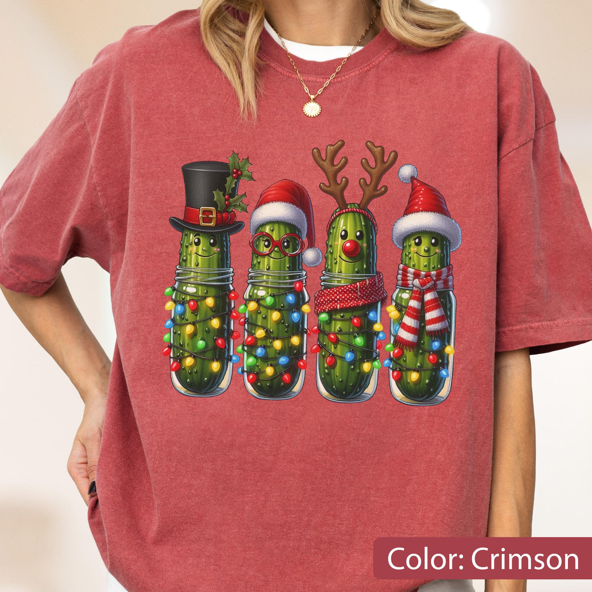 May include: A crimson t-shirt featuring a graphic of four green pickles in glass jars. Each pickle is decorated for the holidays with items including a black top hat, a red Santa hat with glasses, reindeer antlers with a red nose, and a Santa hat with a scarf.