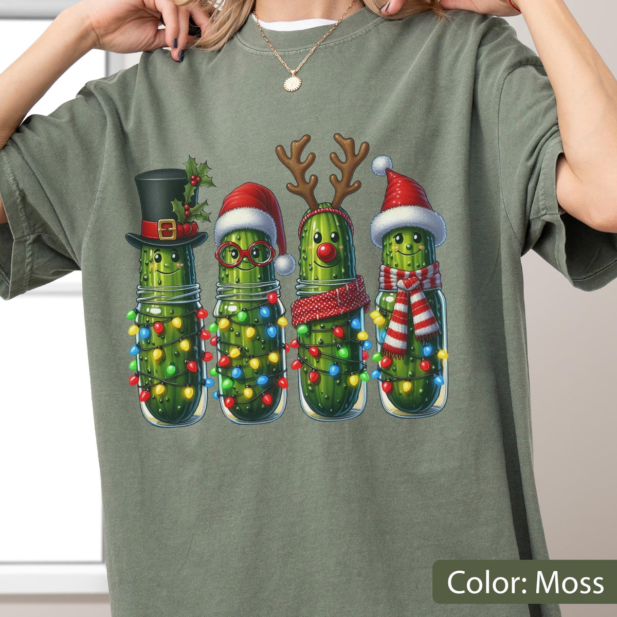 May include: A moss green t-shirt featuring a graphic of four pickles in glass jars. Each pickle is decorated for the holidays with items including a black top hat, a red Santa hat with glasses, reindeer antlers with a red nose, and a Santa hat with a red and white scarf. All jars are wrapped in colorful Christmas lights.