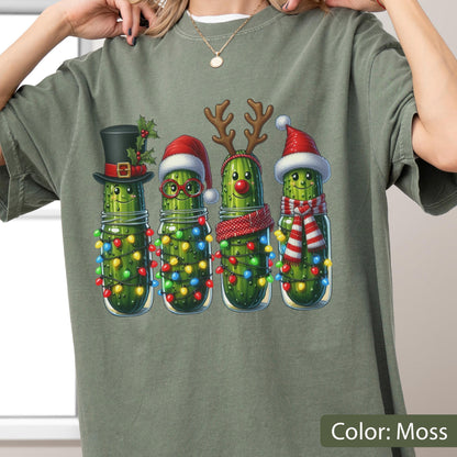 May include: A moss green t-shirt featuring a graphic of four pickles in glass jars. Each pickle is decorated for the holidays with items including a black top hat, a red Santa hat with glasses, reindeer antlers with a red nose, and a Santa hat with a red and white scarf. All jars are wrapped in colorful Christmas lights.