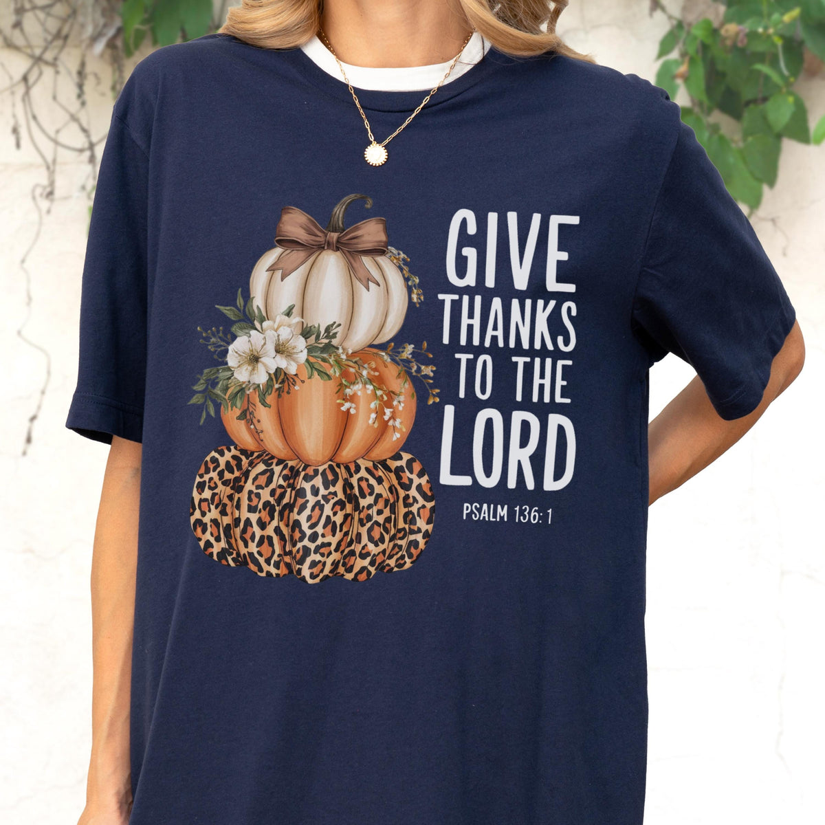 May include: Navy blue t-shirt featuring a stacked pumpkin design with a leopard print base, orange and white pumpkins, and floral accents. The shirt includes the text "GIVE THANKS TO THE LORD" and "PSALM 136:1" in white.