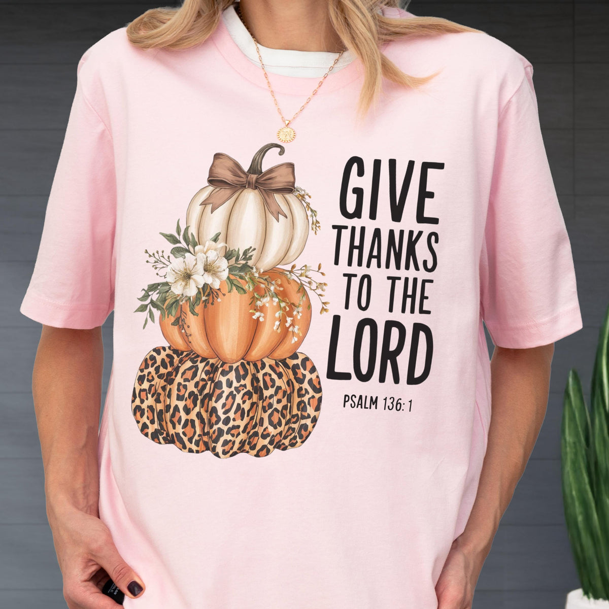 May include: Pink short-sleeved t-shirt featuring a stacked pumpkin design. The pumpkins are white, orange, and leopard print, adorned with floral accents and a brown bow. The text "GIVE THANKS TO THE LORD" is printed on the right side, with "PSALM 136:1" below.