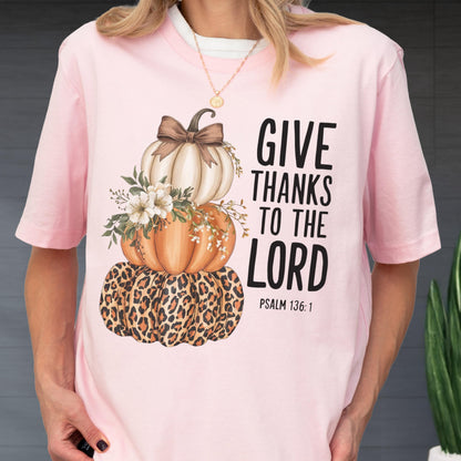 May include: Pink short-sleeved t-shirt featuring a stacked pumpkin design. The pumpkins are white, orange, and leopard print, adorned with floral accents and a brown bow. The text "GIVE THANKS TO THE LORD" is printed on the right side, with "PSALM 136:1" below.