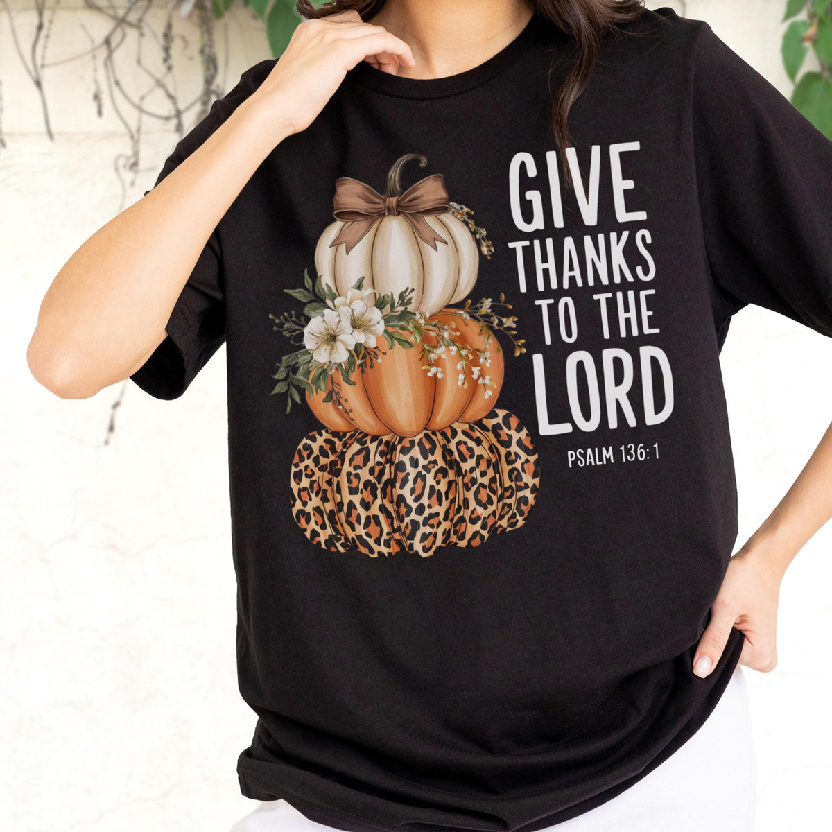 May include: Black t-shirt featuring a stacked pumpkin design with a leopard print base, orange and white pumpkins, and floral accents. The text "GIVE THANKS TO THE LORD" is printed in white, with "PSALM 136:1" below.