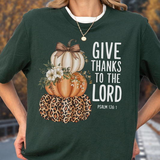 May include: Dark green t-shirt featuring a stacked pumpkin design with a leopard print base, orange and white pumpkins, and floral accents. The text "GIVE THANKS TO THE LORD" is printed in white, with "PSALM 136:1" below.