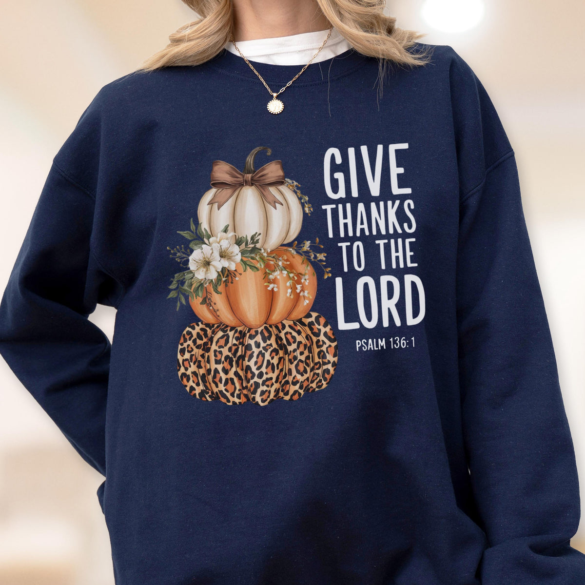 May include: Navy blue sweatshirt featuring a stacked pumpkin design. The pumpkins are white, orange, and leopard print, adorned with flowers and a bow. The text "GIVE THANKS TO THE LORD" is printed in white.