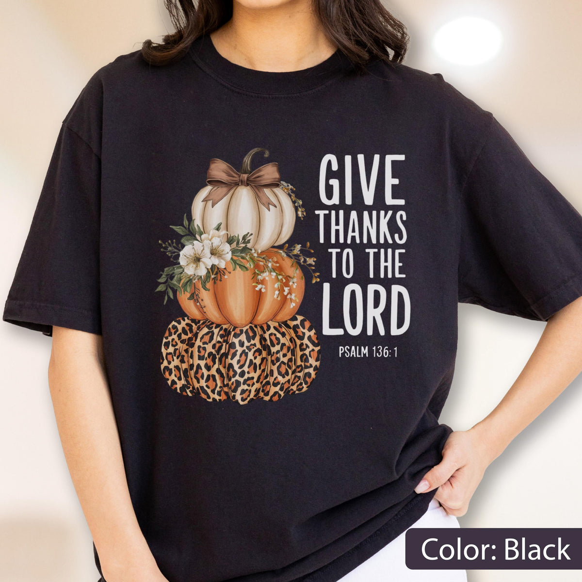 May include: Black t-shirt featuring a stacked pumpkin design. The top pumpkin is white, the middle is orange, and the bottom has a leopard print. The design includes floral accents and the text "GIVE THANKS TO THE LORD" with Psalm 136:1.