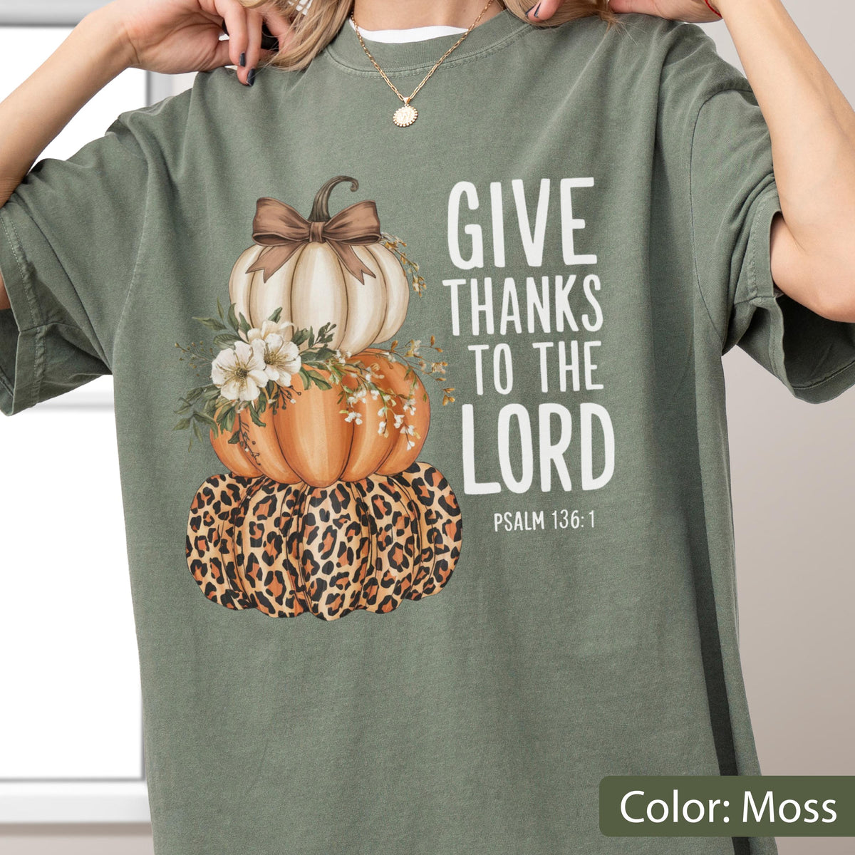 May include: Moss green t-shirt featuring a stacked pumpkin design with a leopard print base, orange and white pumpkins, and a brown bow. The shirt has the text "GIVE THANKS TO THE LORD" and "PSALM 136:1" in white. The shirt is a moss green color.