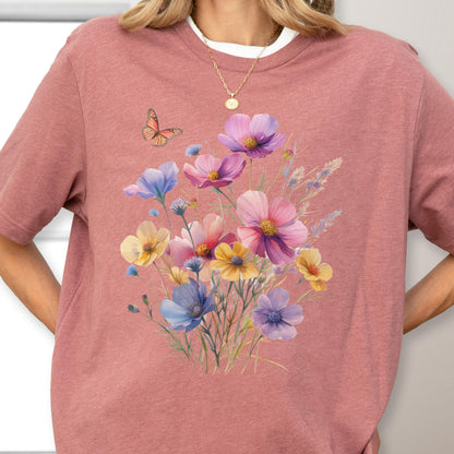 May include: A dusty rose short-sleeved t-shirt featuring a central graphic print of a colorful wildflower bouquet in shades of pink, purple, yellow, and blue, accompanied by a small orange butterfly. The shirt has a crew neckline and a heathered texture.
