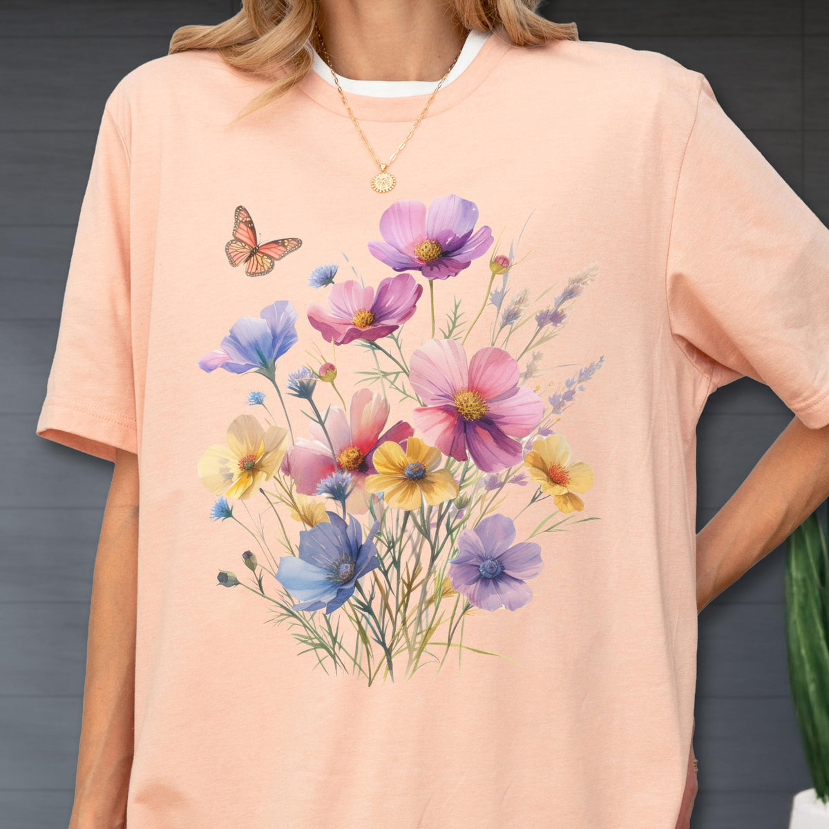 May include: A peach-colored t-shirt featuring a central graphic of a colorful wildflower bouquet in shades of pink, purple, yellow, and blue, accompanied by a single orange butterfly. The design is printed on the front of the short-sleeved garment.