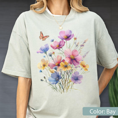 May include: A light sage green t-shirt featuring a central graphic of a colorful wildflower bouquet in shades of pink, yellow, blue, and purple, accompanied by a single orange butterfly. The shirt has a crew neckline and short sleeves.
