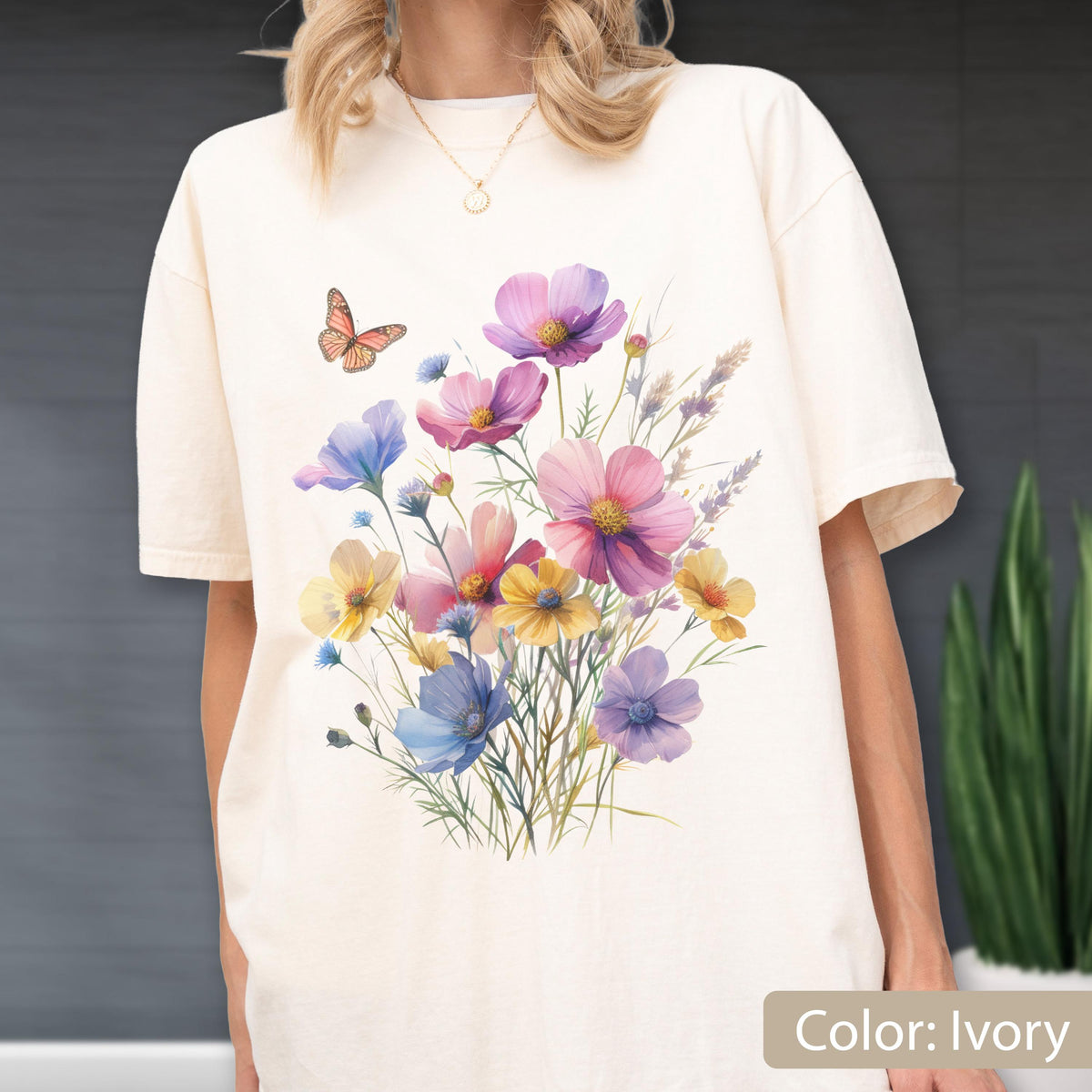May include: An ivory short-sleeve t-shirt featuring a colorful graphic print of a wildflower bouquet with pink, yellow, and blue blossoms, green stems, and a single orange butterfly. The shirt has a crew neckline and a relaxed, loose fit.