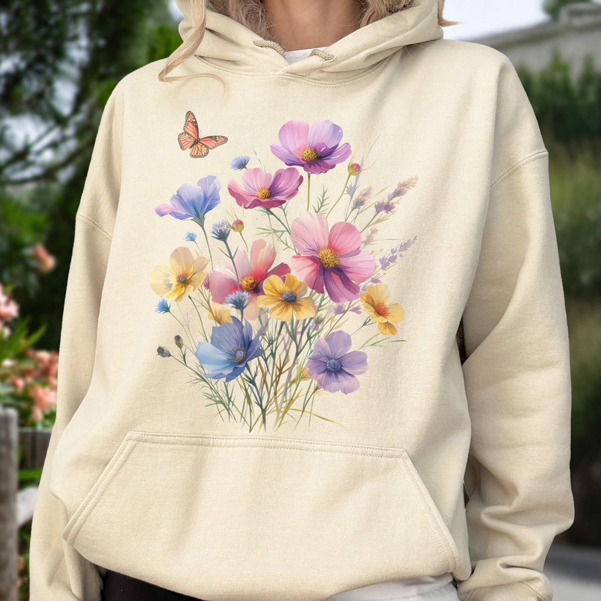 May include: Beige hoodie featuring a watercolor floral design. The design includes a bouquet of colorful flowers in shades of pink, purple, yellow, and blue, with a small orange butterfly. The hoodie has a front pocket and a drawstring hood.