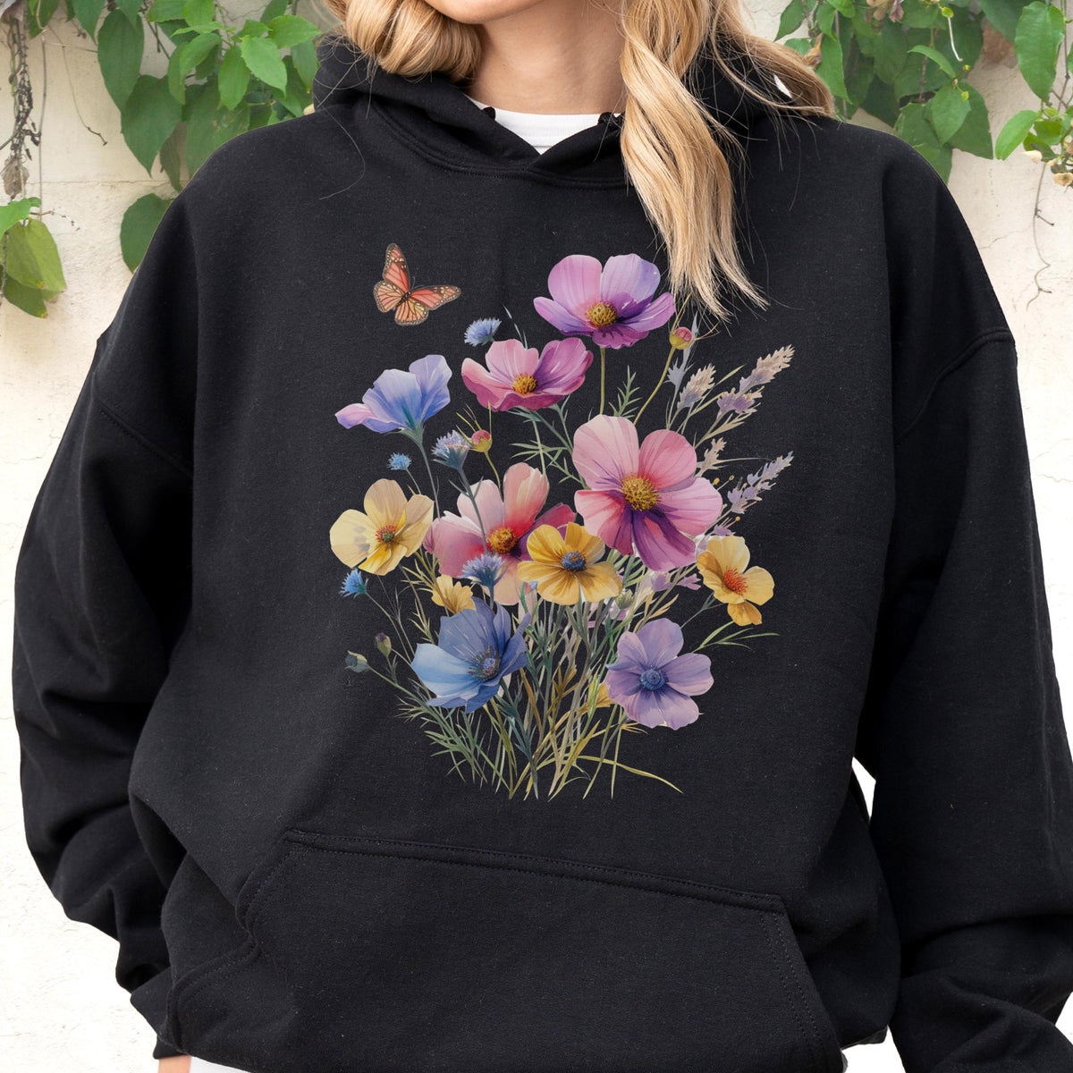 May include: Black hooded sweatshirt featuring a watercolor floral design with pink, purple, yellow, and blue flowers. A small orange butterfly is also present. The sweatshirt has a front pocket.