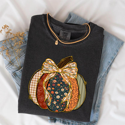 May include: Dark gray Comfort Colors t-shirt featuring a colorful pumpkin design. The pumpkin has a plaid, floral, and leopard print pattern with a large bow. A gold necklace is draped over the shirt, which is folded on top of blue jeans.
