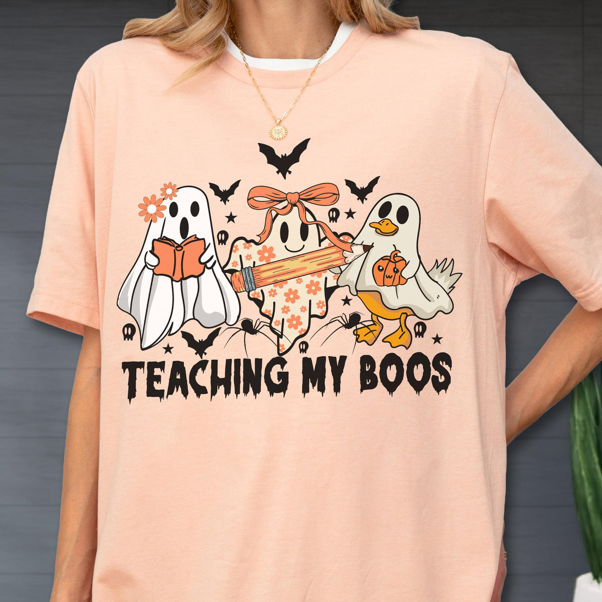 May include: Peach-colored t-shirt with a Halloween-themed graphic. The design features three cartoon ghosts, bats, spiders, and the text "TEACHING MY BOOS". One ghost holds a book, another a pencil, and the third a pumpkin.