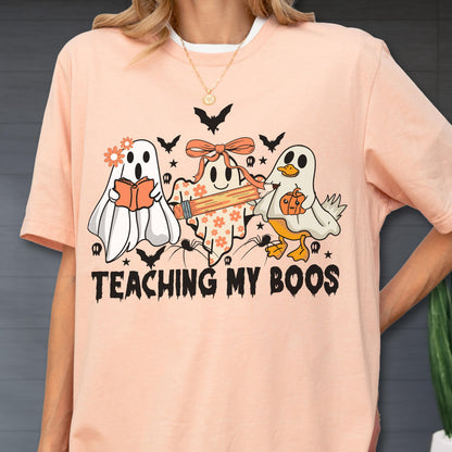 May include: Peach-colored t-shirt with a Halloween-themed graphic. The design features three cartoon ghosts, bats, spiders, and the text "TEACHING MY BOOS". One ghost holds a book, another a pencil, and the third a pumpkin.