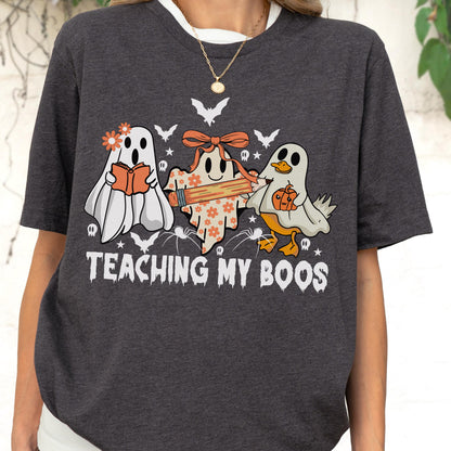 May include: Dark gray t-shirt with a white collar featuring a Halloween-themed graphic. The graphic includes three cartoon ghosts, bats, spiders, and the text "TEACHING MY BOOS". The ghosts are holding a book, a pencil, and a pumpkin.