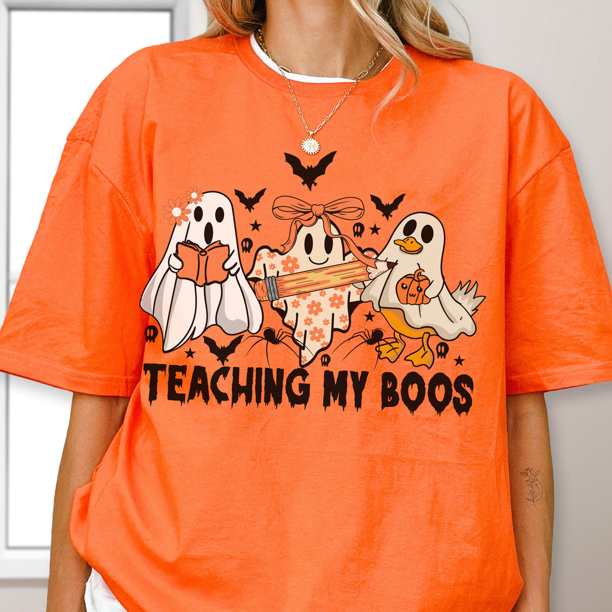 May include: Orange t-shirt with a Halloween-themed graphic. The design features three cartoon ghosts, bats, spiders, and the text "TEACHING MY BOOS". One ghost reads a book, another holds a pencil, and the third is a duck holding a pumpkin.