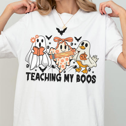 May include: White t-shirt with a Halloween-themed graphic. The design features three cartoon ghosts, one reading a book, one holding a pencil, and one with a pumpkin. The text "TEACHING MY BOOS" is printed below the ghosts.