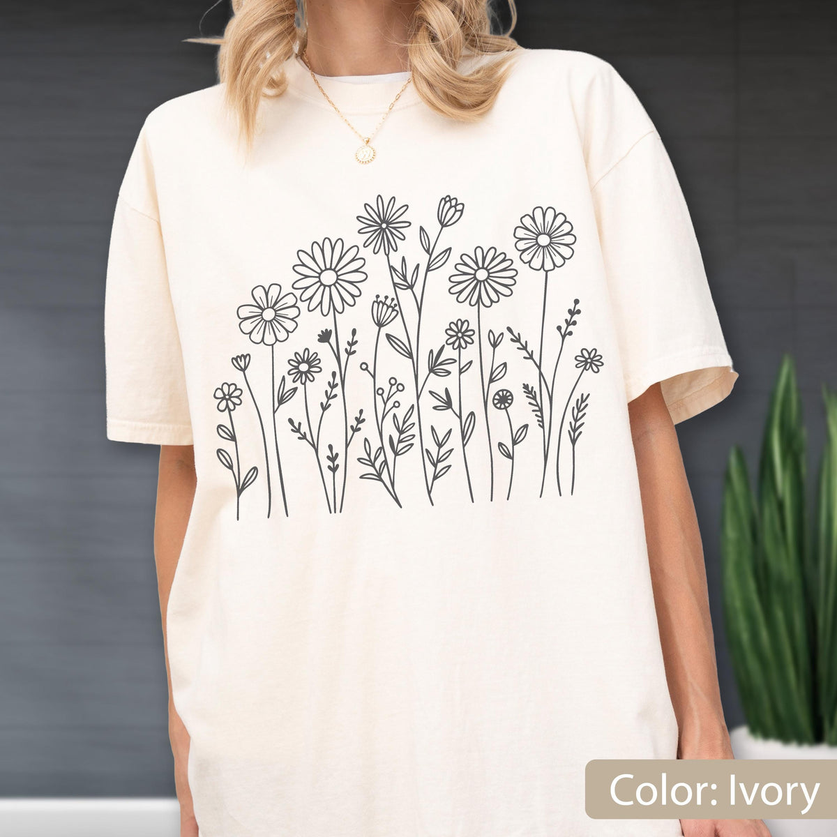 May include: An ivory t-shirt featuring a black line art graphic of various wildflowers and daisies across the front. The shirt has a crew neckline and short sleeves, showcasing a simple botanical illustration of stems, leaves, and flower heads.