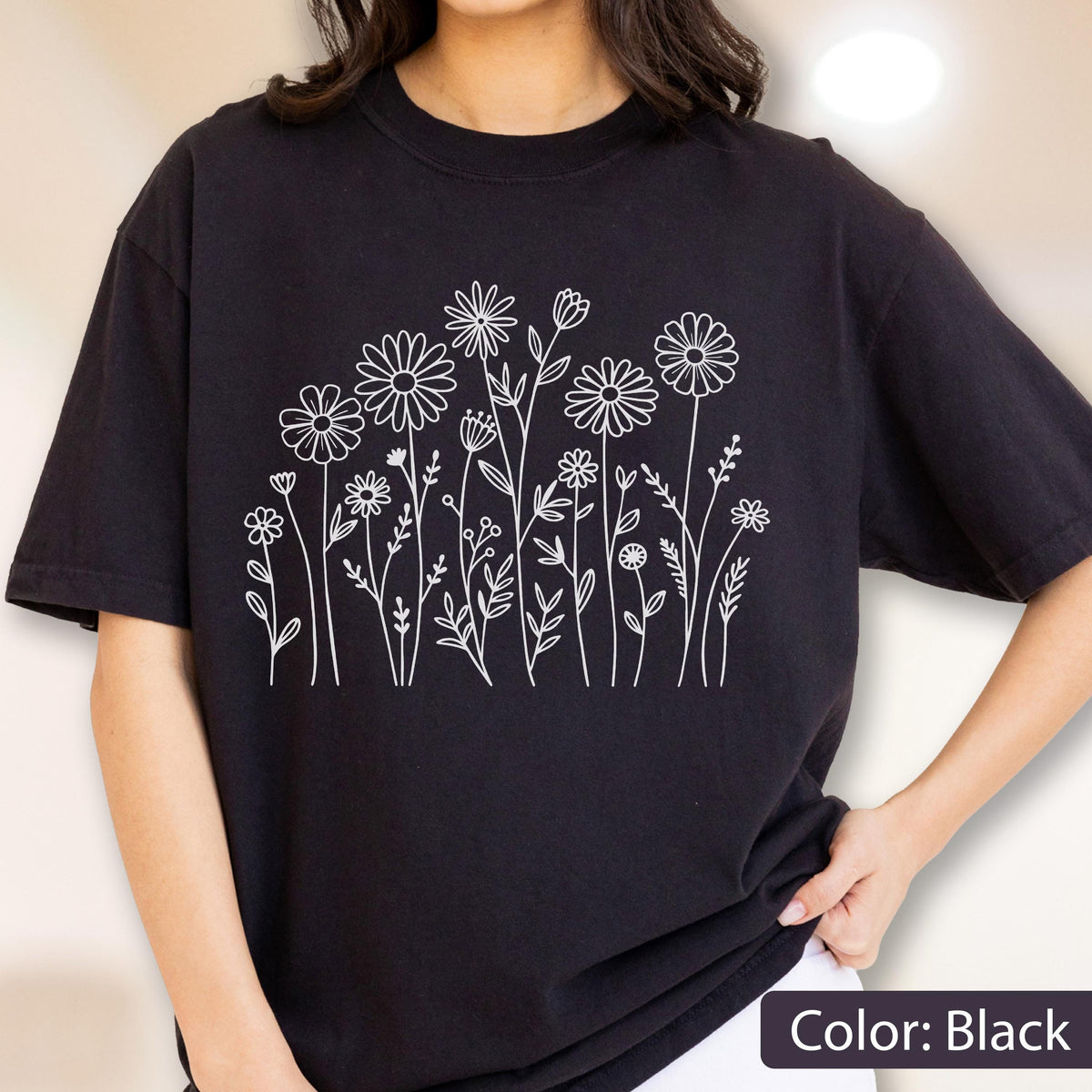 May include: A black short-sleeved t-shirt featuring a white line art graphic of various wildflowers and stems across the chest. The design consists of multiple daisy-like flowers and thin botanical elements arranged in a cluster on the front.