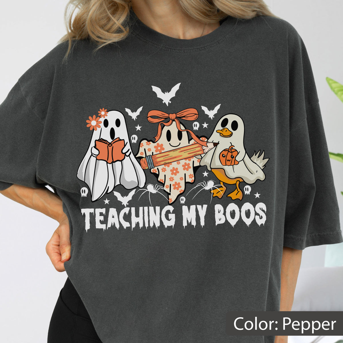 May include: Pepper-colored t-shirt with a Halloween-themed graphic. The design features three cartoon ghosts, one reading a book, one holding a pencil, and one with a pumpkin. The text "TEACHING MY BOOS" is printed below the ghosts.