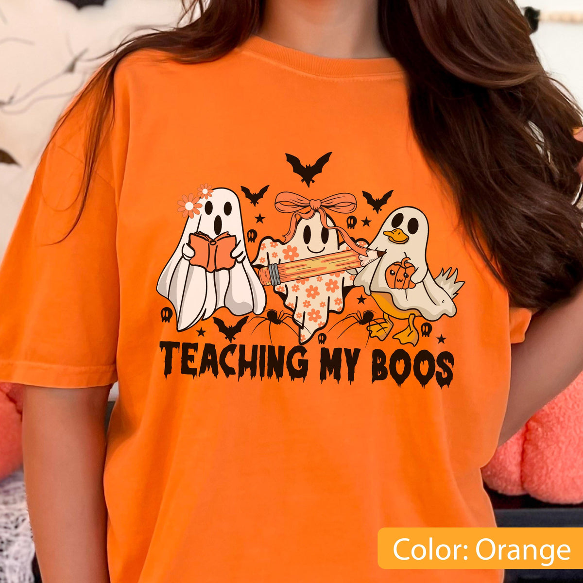 May include: Orange t-shirt with a Halloween-themed graphic. The graphic features three cartoon ghosts, bats, and spiders, with the text "TEACHING MY BOOS". One ghost reads a book, another holds a pencil, and the third holds a pumpkin.