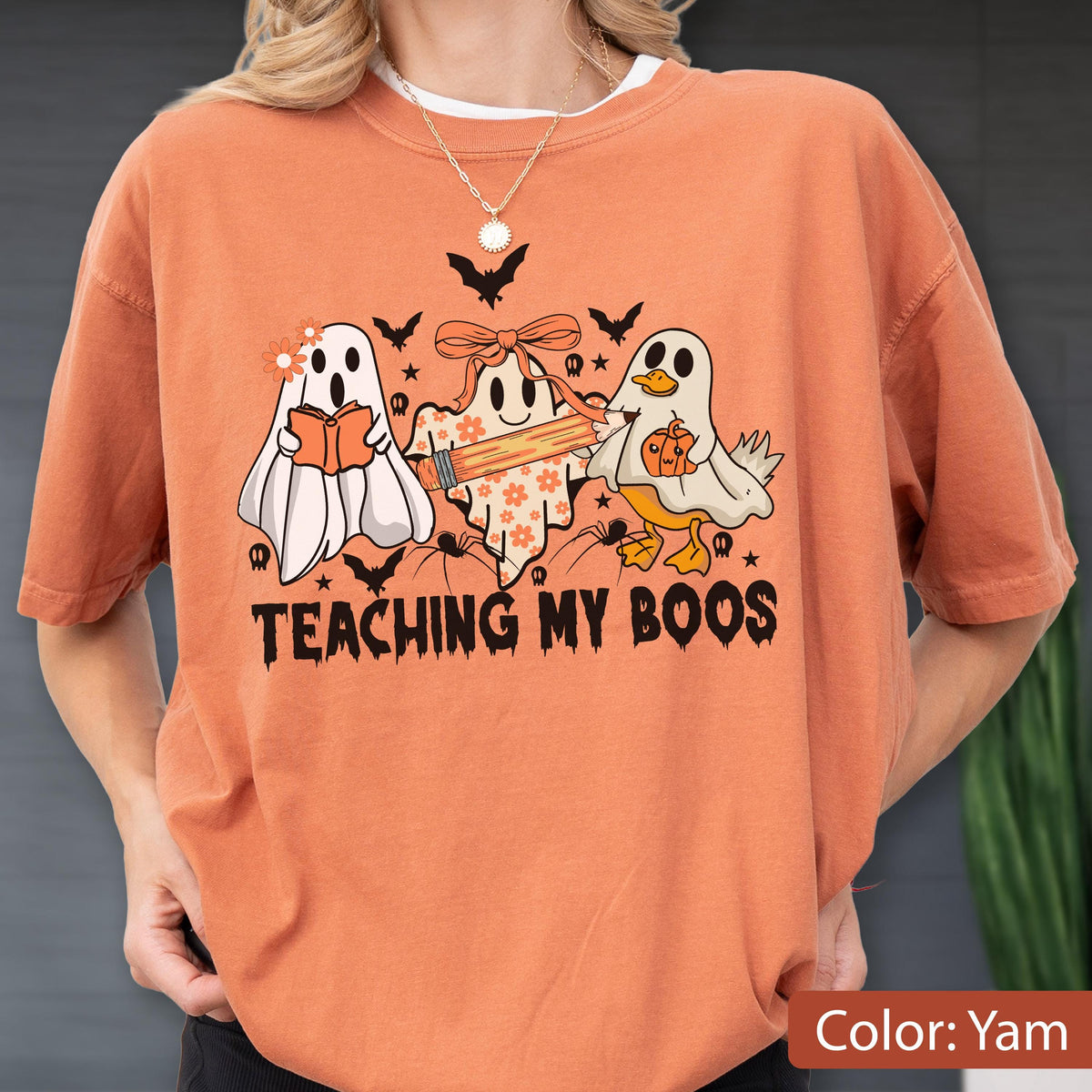 May include: Orange-colored t-shirt with a Halloween-themed graphic. The design features three cartoon ghosts, bats, and the text "TEACHING MY BOOS". One ghost reads a book, another holds a pencil, and the third holds a pumpkin.