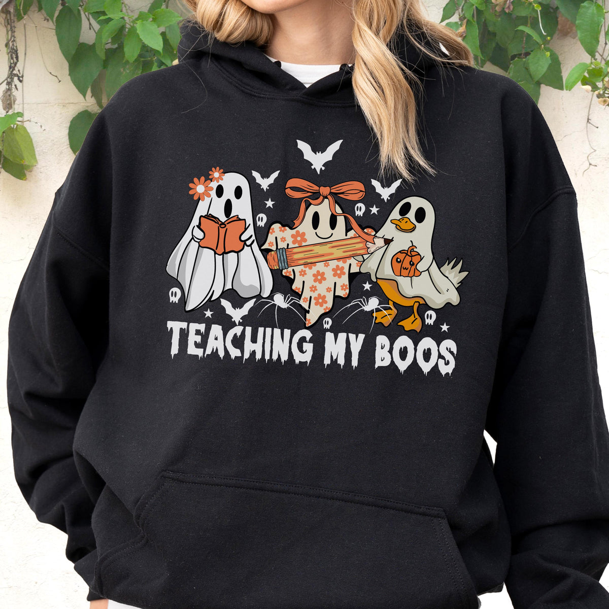 May include: A black hooded sweatshirt featuring a graphic of three ghosts, one holding a book, one with a pencil, and one with a pumpkin. The design includes small bats, spiders, and flowers, with the text "TEACHING MY BOOS" printed in white.