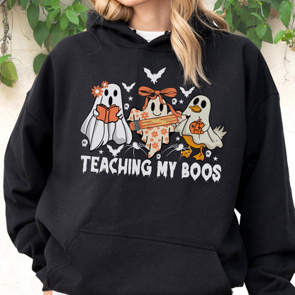 May include: A black hooded sweatshirt featuring a graphic of three ghosts, one holding a book, one with a pencil, and one with a pumpkin. The design includes small bats, spiders, and flowers, with the text "TEACHING MY BOOS" printed in white.