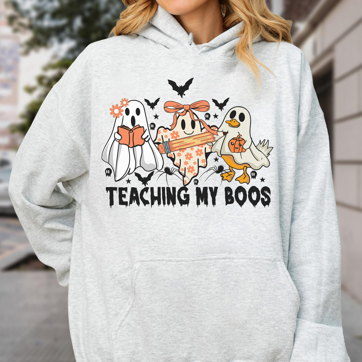 May include: A light gray hooded sweatshirt featuring a graphic print of three ghosts, one holding a book, one with a pencil, and one holding a pumpkin. The design includes small bats, spiders, and stars with the text "TEACHING MY BOOS" in black.