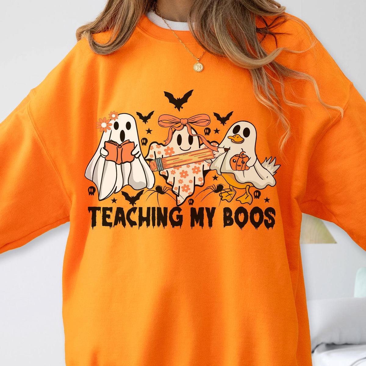 May include: An orange crewneck sweatshirt featuring a graphic of three ghosts, one holding a book, one holding a pencil, and one holding a pumpkin. The design includes bats, spiders, and the text "TEACHING MY BOOS" in a black, dripping font.