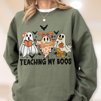 May include: An olive green crewneck sweatshirt featuring a graphic of three ghosts. One ghost holds a book, one holds a pencil, and one is a ghost duck holding a pumpkin. The text "TEACHING MY BOOS" is printed in black below the ghosts with bats.
