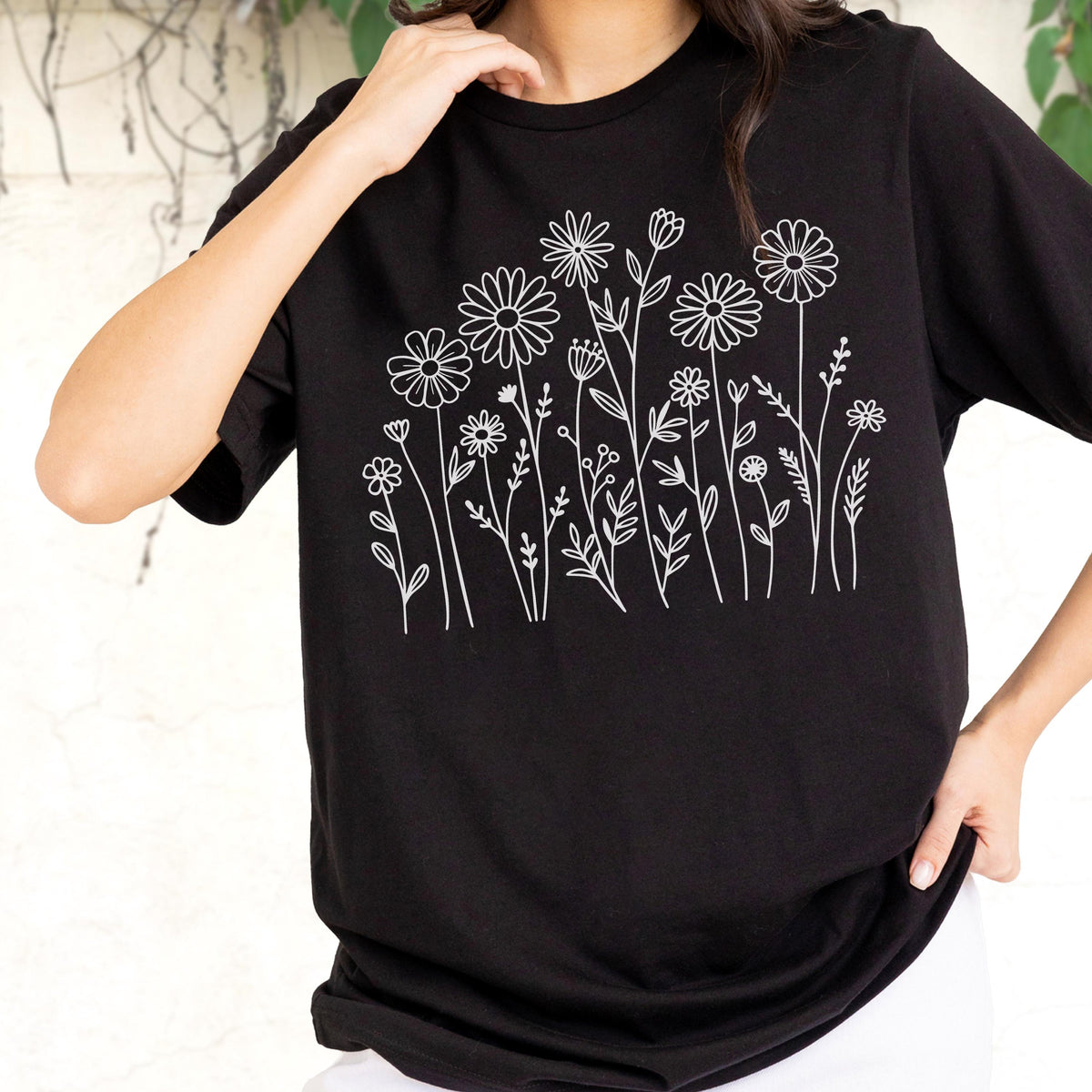 May include: A black short-sleeved t-shirt featuring a white line art graphic of various wildflowers and daisies across the chest. The design consists of thin, clean outlines of different floral stems, leaves, and blossoms arranged in a cluster.