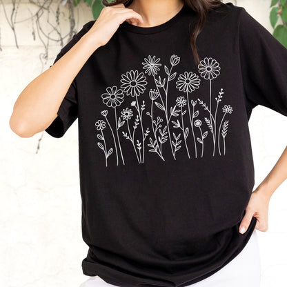 May include: A black short-sleeved t-shirt featuring a white line art graphic of various wildflowers and daisies across the chest. The design consists of thin, clean outlines of different floral stems, leaves, and blossoms arranged in a cluster.