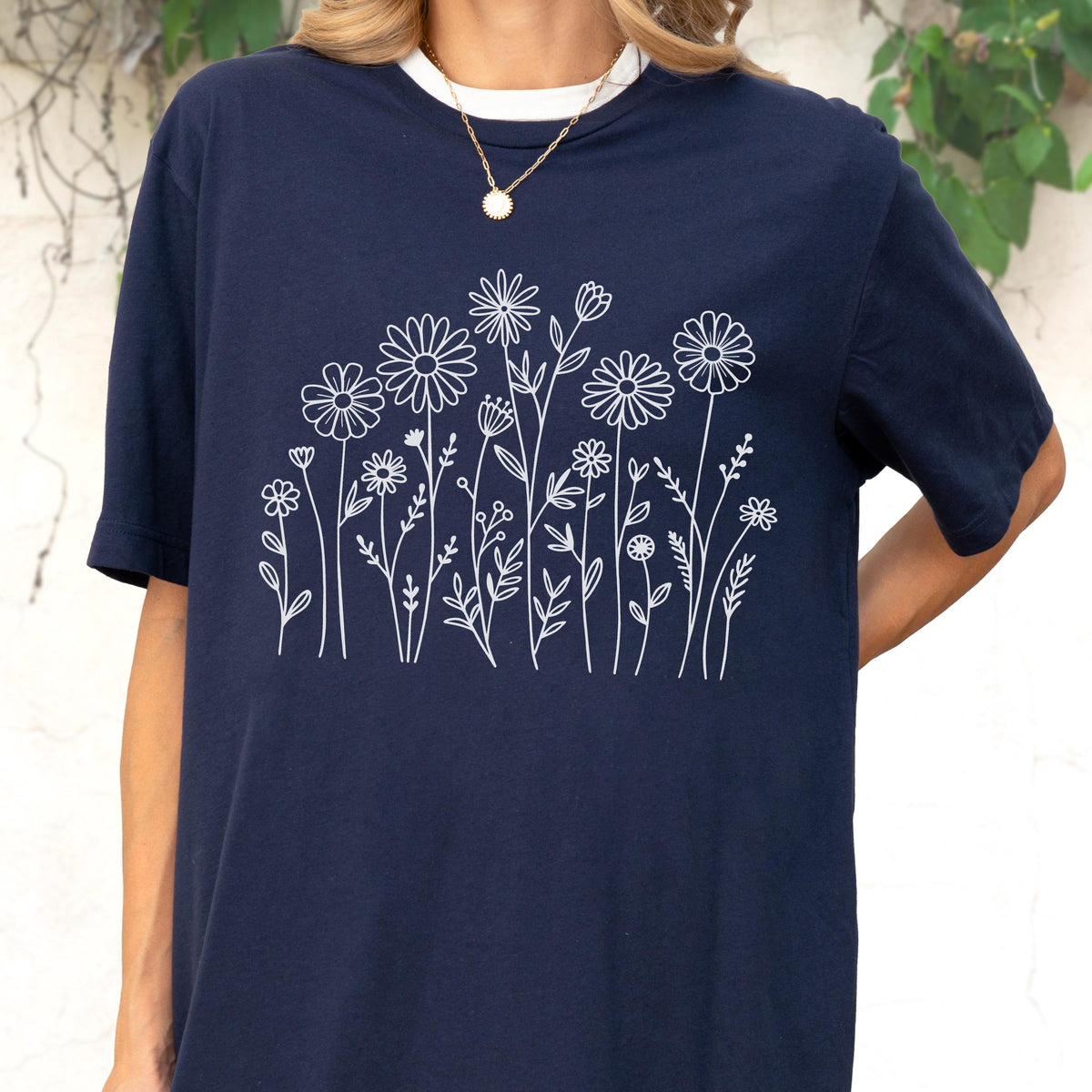 May include: A navy blue short-sleeve t-shirt featuring a white line art graphic of various wildflowers and daisies across the chest. The shirt has a crew neckline and is worn with a thin gold chain necklace over a white undershirt layer.
