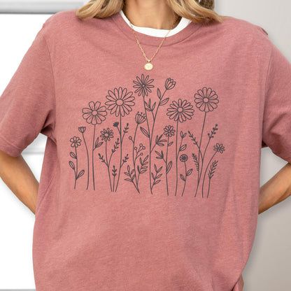 May include: A mauve short-sleeved t-shirt featuring a black line art graphic of various wildflowers and stems across the chest. The shirt has a crew neckline and is worn with a thin gold chain necklace with a small circular pendant.