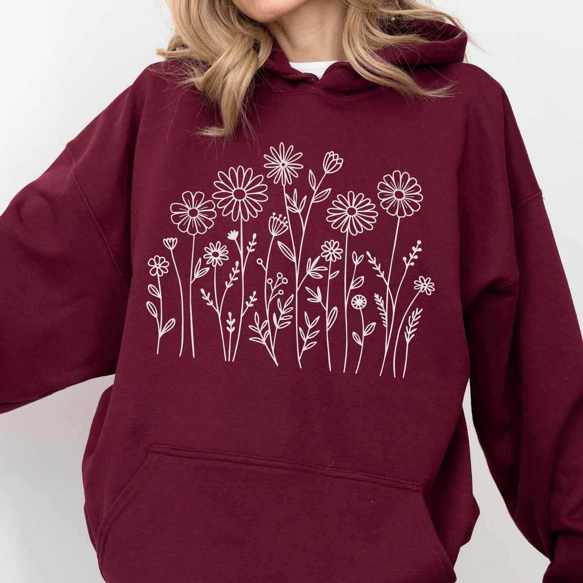 May include: A maroon hooded sweatshirt featuring a white line art graphic of various wildflowers and stems across the chest. The garment includes a hood with drawstrings and a large front kangaroo pocket, showcasing a simple floral design.