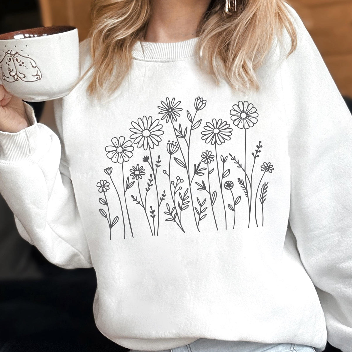 May include: A white crewneck sweatshirt featuring a black line art illustration of various wildflowers and daisies centered on the chest. The design consists of multiple stems, leaves, and flower heads arranged in a botanical pattern on the fabric.