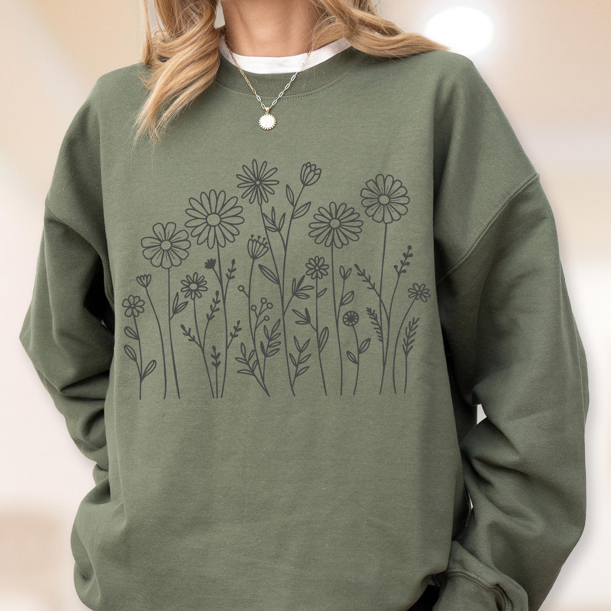 May include: An olive green crewneck sweatshirt featuring a dark gray line art illustration of various wildflowers and daisies across the chest. The garment has long sleeves and a ribbed neckline, worn with a simple silver chain necklace.