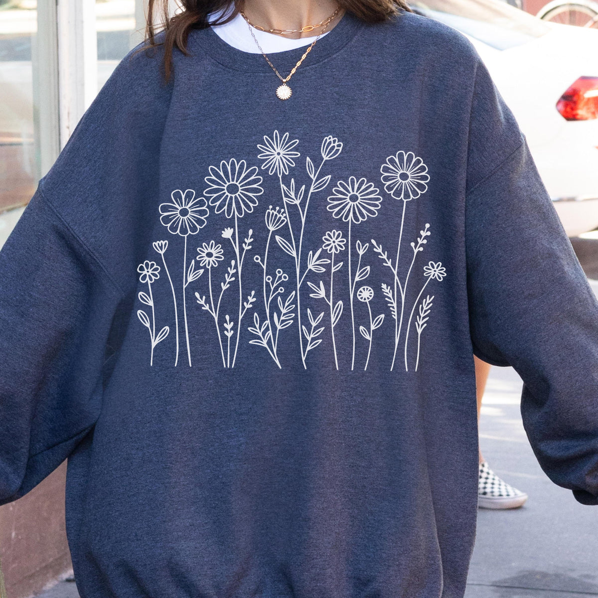 May include: A dark heather navy blue crewneck sweatshirt featuring a white line art graphic of various wildflowers and stems across the chest. The long-sleeved garment has a ribbed collar, cuffs, and hem, showcasing a simple botanical illustration.