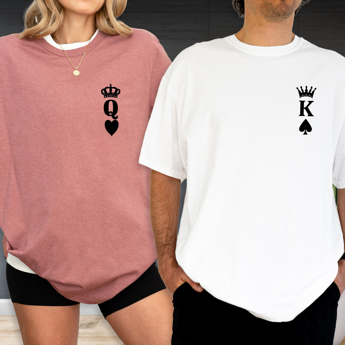May include: A pair of matching shirts, one dusty rose and one white. The dusty rose shirt features a black crown, the letter "Q", and a heart. The white shirt has a black crown, the letter "K", and a spade. The shirts are casual wear.