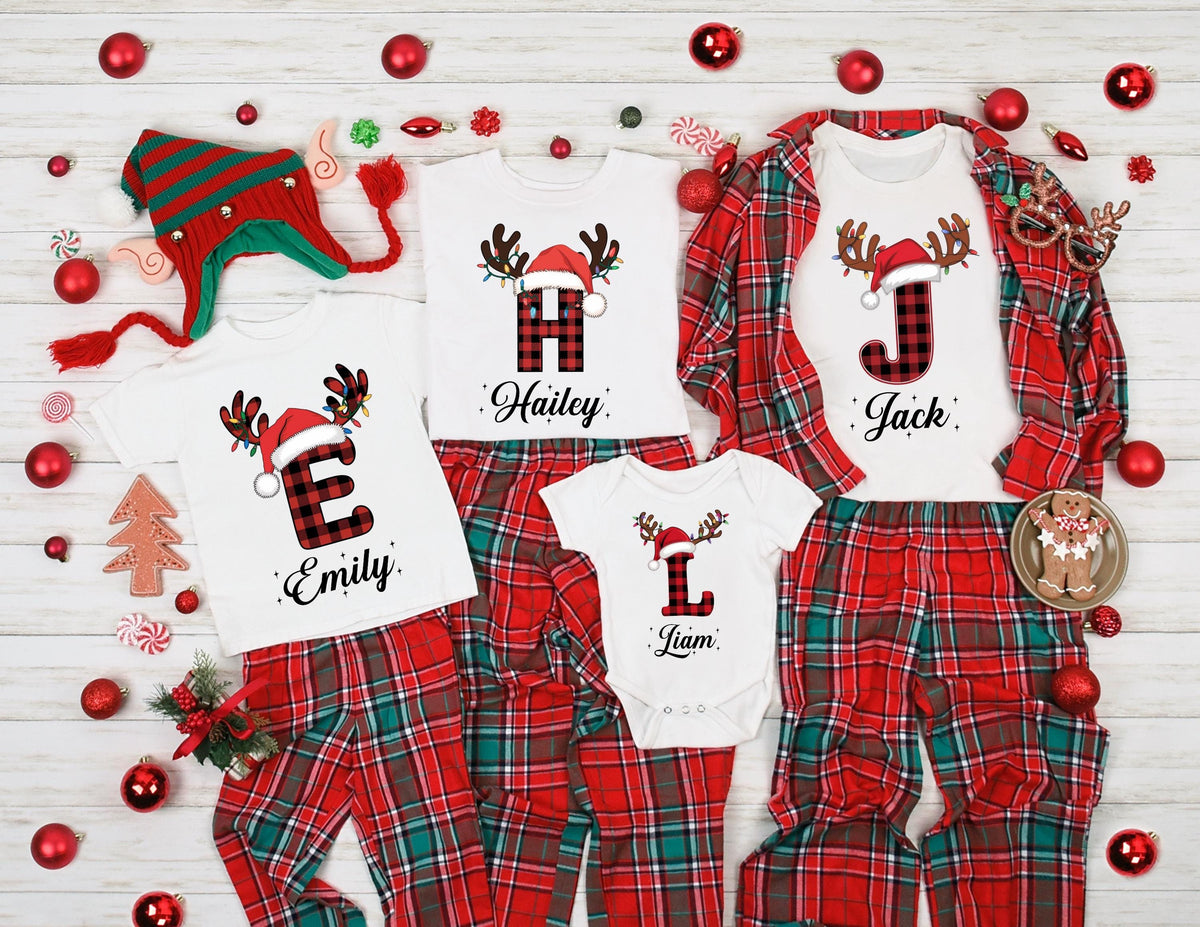 May include: Matching family Christmas pajamas in red and green plaid. White shirts with reindeer antlers, Santa hats, and initial letters. The names "Emily", "Hailey", "Jack", and "Liam" are printed below the letters. A Christmas hat and ornaments complete the festive scene.