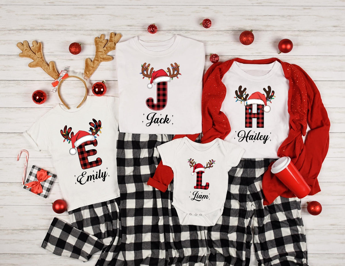 May include: Matching family Christmas pajamas with reindeer antlers and Santa hats. The shirts display names and initial letters in a plaid pattern. The set includes a headband, red ornaments, and black and white plaid pants.