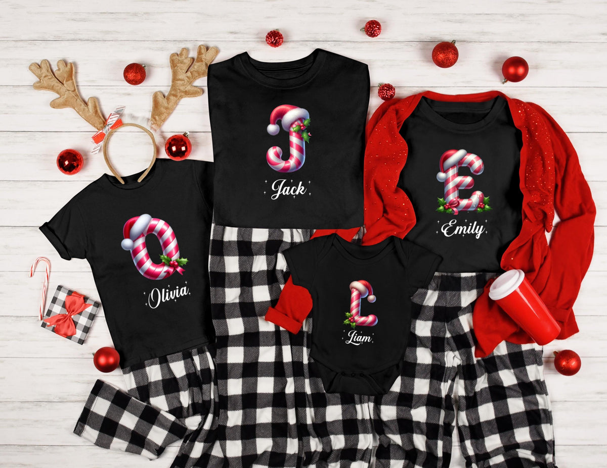 May include: Black t-shirts and a onesie with candy cane initial designs and names, paired with black and white plaid pajama pants. Festive Christmas apparel with Santa hats and holiday decorations.