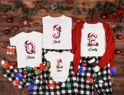 May include: White Christmas-themed shirts with names and candy cane letter designs. The shirts are paired with black and white plaid pajama pants. Festive holiday decor includes ornaments, lights, and greenery.