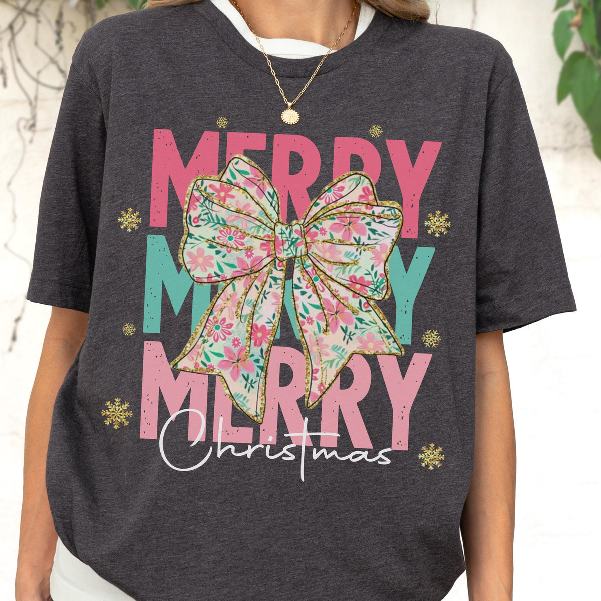 May include: Dark gray t-shirt with the words "MERRY MERRY MERRY Christmas" in pink and teal. A large floral bow is centered on the shirt. Gold snowflakes are scattered around the text.