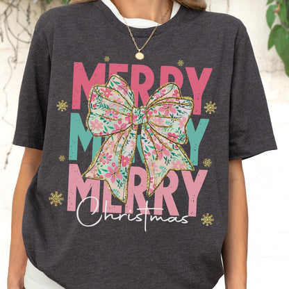 May include: Dark gray t-shirt with the words "MERRY MERRY MERRY Christmas" in pink and teal. A large floral bow is centered on the shirt. Gold snowflakes are scattered around the text.